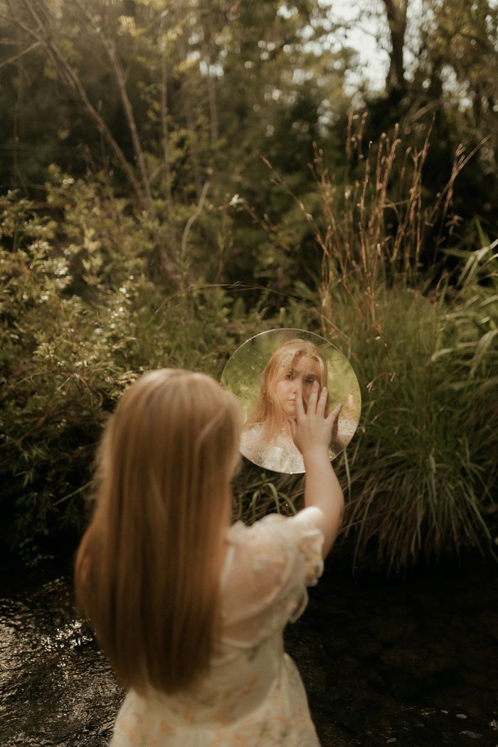 who-we-are A young girl observes her reflection in a mirror outdoors, surrounded by lush nature.